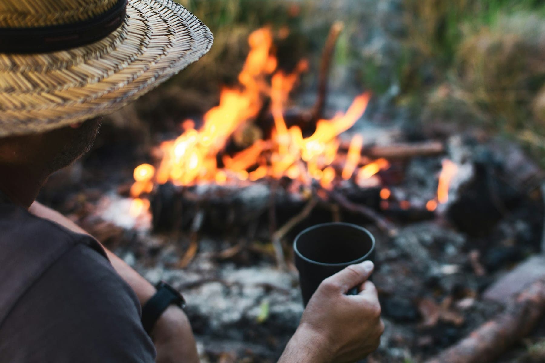 A person in a straw hat holding a tin mug beside a campfire